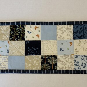 May include: A rectangular quilted table runner featuring a patchwork design of various fabric squares in shades of navy blue, light blue, cream, and white. The patterns include botanical prints, small birds, and script text, finished with a dark blue striped border.