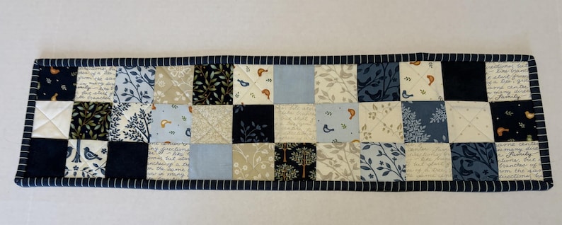 May include: A rectangular quilted table runner featuring a patchwork design of various fabric squares in shades of navy blue, light blue, cream, and white. The patterns include botanical prints, small birds, and script text, finished with a dark blue striped border.