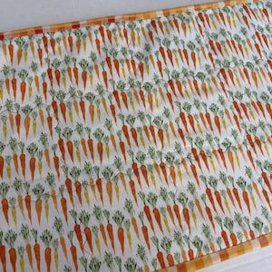 May include: A rectangular quilted table runner featuring a repeating pattern of orange carrots with green leafy tops on a white background, finished with a decorative orange and white checkered fabric border along the edges.