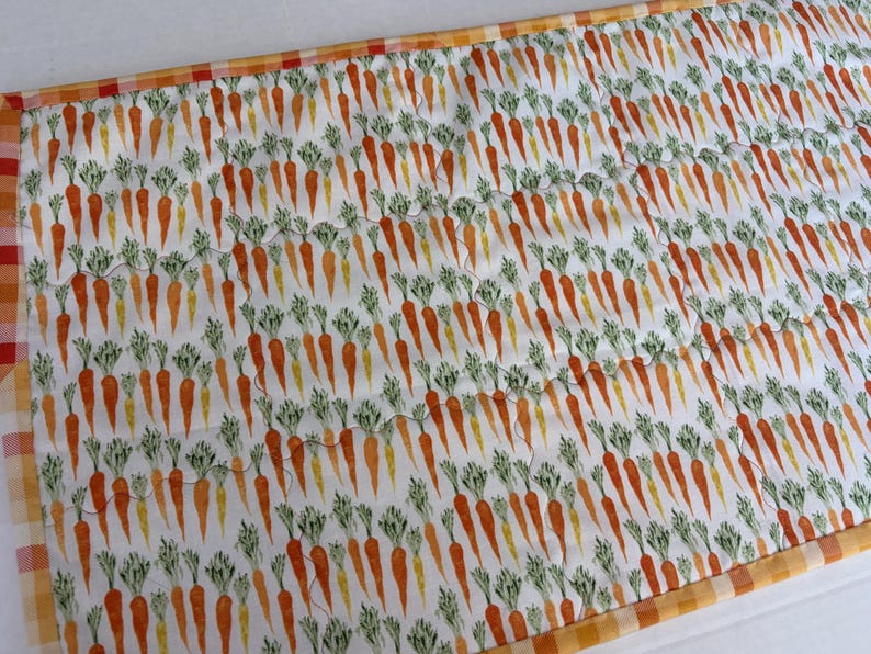 May include: A rectangular quilted table runner featuring a repeating pattern of orange carrots with green leafy tops on a white background, finished with a decorative orange and white checkered fabric border along the edges.