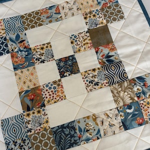 May include: A square quilted fabric piece featuring a patchwork design of various floral and geometric patterns in shades of blue, cream, tan, and orange. The quilt is constructed with alternating solid cream squares and patterned squares, finished with a dark blue border and diamond-shaped machine quilting stitches.