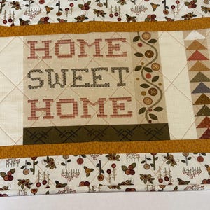 May include: A quilted table runner featuring the text HOME SWEET HOME in brown cross-stitch style lettering. The fabric displays a cream background with patterns of small flowers, berries, geometric triangles, and house motifs in mustard, brown, and red.
