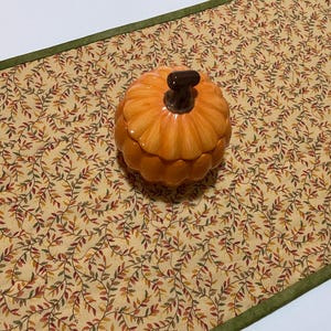 Puede incluir: Un recipiente de cer&aacute;mica naranja con forma de calabaza y un tallo marr&oacute;n oscuro se encuentra sobre un camino de mesa beige. El camino de mesa tiene un patr&oacute;n repetido de hojas verdes, rojas y marrones. Un borde verde enmarca el camino de mesa.