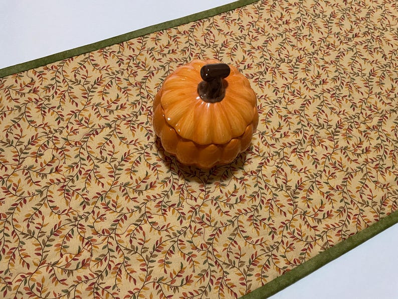 Puede incluir: Un recipiente de cer&aacute;mica naranja con forma de calabaza y un tallo marr&oacute;n oscuro se encuentra sobre un camino de mesa beige. El camino de mesa tiene un patr&oacute;n repetido de hojas verdes, rojas y marrones. Un borde verde enmarca el camino de mesa.