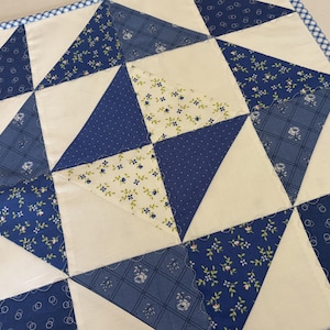 May include: A square fabric quilt featuring a geometric pattern of alternating cream and blue triangles. The blue fabric sections display various patterns including floral prints, plaid, and circles. The quilt is finished with a blue and white gingham border.