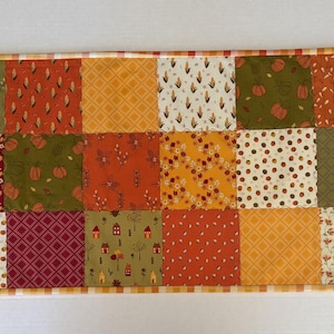 May include: A rectangular patchwork table runner featuring a grid of fabric squares in autumn colours including orange, olive green, burgundy, and cream. The patterns include pumpkins, small houses, floral motifs, geometric shapes, and leaves.