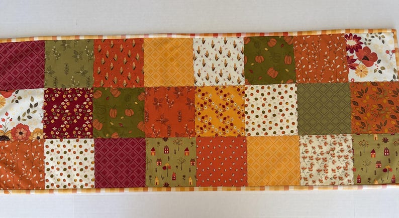 May include: A rectangular patchwork table runner featuring a grid of fabric squares in autumn colours including orange, olive green, burgundy, and cream. The patterns include pumpkins, small houses, floral motifs, geometric shapes, and leaves.