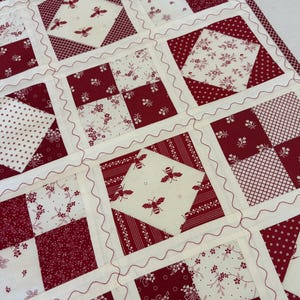 May include: A patchwork quilt featuring a repeating grid of square blocks in deep red and cream. The design includes alternating patterns of floral prints, small dots, cross-hatching, and bee motifs. Each block is separated by cream sashing with red wavy decorative stitching.