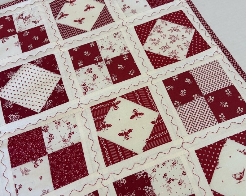 May include: A patchwork quilt featuring a repeating grid of square blocks in deep red and cream. The design includes alternating patterns of floral prints, small dots, cross-hatching, and bee motifs. Each block is separated by cream sashing with red wavy decorative stitching.