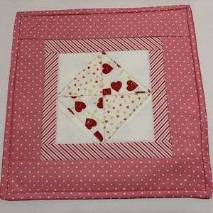 May include: A square, quilted hot pad with a pink and white heart pattern. The center features a white diamond shape with red heart accents. The border is pink with white polka dots and red and white stripes.
