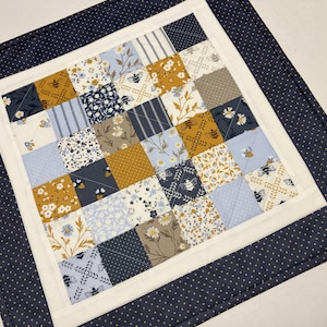 May include: A handmade patchwork quilt with a square design. The center features a grid of small squares in various patterns, including floral, bee, and striped designs in shades of blue, gold, and white. The quilt is bordered by a white frame and a navy blue polka dot fabric.