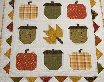 May include: A quilted autumn-themed wall hanging featuring appliqued pumpkins, acorns, and a leaf. The central design is on a white background, with a border of triangles in fall colors. The quilt is machine-stitched.