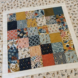 May include: A square patchwork quilt with a white border and a bird-patterned fabric edge. The center features a grid of colorful squares with floral, geometric, and bird designs in shades of blue, orange, yellow, and cream.