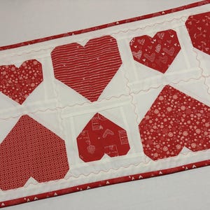 May include: A rectangular quilted table runner featuring a repeating pattern of red hearts on a white background. The hearts have various patterns, including stripes and polka dots. The edges are finished with a red border.