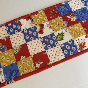 May include: A rectangular quilted table runner featuring a patchwork design of squares and triangles. The fabric pattern includes floral and leaf motifs in shades of red, mustard yellow, cream, and blue, with visible diagonal stitching throughout.