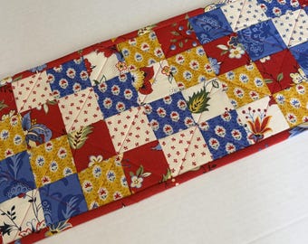 May include: A rectangular quilted table runner featuring a patchwork design of squares and triangles. The fabric pattern includes floral and leaf motifs in shades of red, mustard yellow, cream, and blue, with visible diagonal stitching throughout.