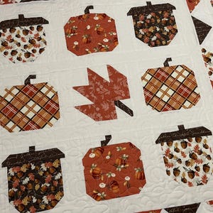 May include: A quilted autumn-themed blanket featuring appliqued pumpkins, acorns, and a leaf. The pumpkins and acorns have brown stems and are made from various patterned fabrics in orange, brown, and white. The leaf is a solid orange fabric. The background is white.
