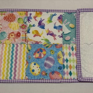 Może przedstawiać: Pikowana podkładka wielkanocna z patchworkowym wzorem. Tkanina przedstawia kolorowe wzory pisanek, kr&oacute;liczk&oacute;w i motyw&oacute;w kwiatowych. Krawędzie obszyte są fioletowo-białą tkaniną w kratkę.