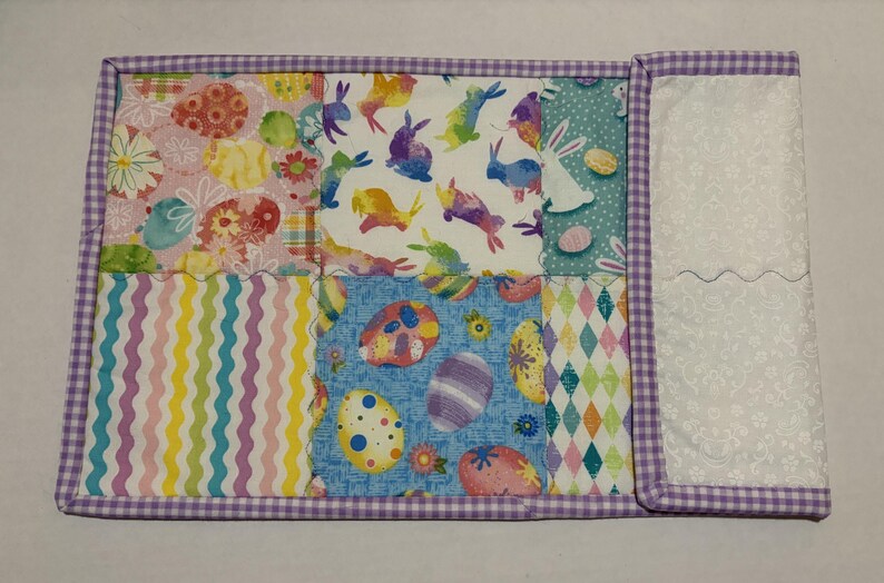 Może przedstawiać: Pikowana podkładka wielkanocna z patchworkowym wzorem. Tkanina przedstawia kolorowe wzory pisanek, kr&oacute;liczk&oacute;w i motyw&oacute;w kwiatowych. Krawędzie obszyte są fioletowo-białą tkaniną w kratkę.