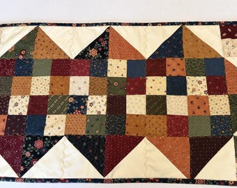 May include: A rectangular table runner featuring a patchwork design of small squares in shades of red, blue, green, and cream. The fabric pieces are arranged in a geometric pattern with large cream triangles and a dark floral border trim.