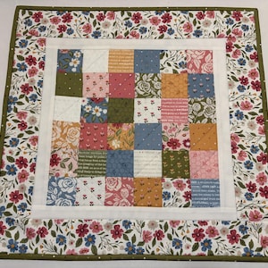 May include: A square patchwork quilt with a floral border. The center features a grid of small squares in various floral patterns and colors, including pink, blue, and gold. The border is white with a green edge, and the outer edge has a floral pattern.