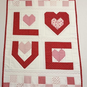 May include: A quilted wall hanging with the word "LOVE" spelled out in red fabric. Each letter features a heart design with various patterns, including stripes, polka dots, and floral prints. The background is white with a red polka dot border.