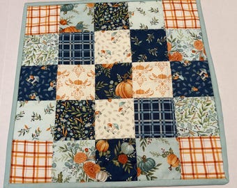 May include: A square patchwork quilt featuring a grid of twenty-five fabric squares with various patterns including orange plaid, navy blue plaid, pumpkins, mushrooms, and floral motifs, all bordered by a solid light blue fabric binding edge.