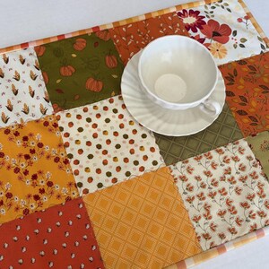 May include: A rectangular patchwork table runner featuring various autumn-themed fabric squares in shades of orange, yellow, green, and white, with patterns including pumpkins, corn, flowers, and houses, topped with a white ceramic teacup and saucer.