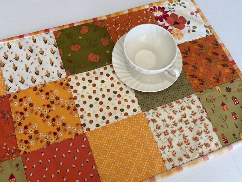 May include: A rectangular patchwork table runner featuring various autumn-themed fabric squares in shades of orange, yellow, green, and white, with patterns including pumpkins, corn, flowers, and houses, topped with a white ceramic teacup and saucer.