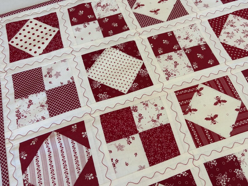 May include: A patchwork quilt featuring a repeating grid of square blocks in deep red and cream. The design alternates between four-patch floral patterns, polka dots, stripes, and cream squares printed with small red bees, all joined by wavy stitching.