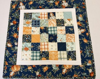 May include: A square fabric quilt featuring a central patchwork grid of small squares in shades of navy blue, orange, cream, and sage green. The patterns include pumpkins, plaid, and foliage. A white border frames the grid, surrounded by a navy blue border.