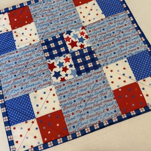 May include: A square quilted placemat with a patriotic theme. It features red, white, and blue squares with star patterns and the text "Sweet Home" and "America". The border is a blue and white checkered pattern.