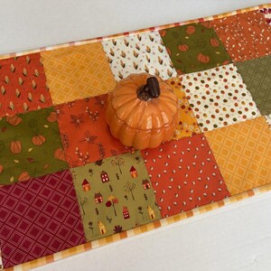 May include: A rectangular patchwork table runner featuring a variety of autumn-themed fabric squares in shades of orange, green, red, and cream, topped with a small, glossy, orange ceramic pumpkin-shaped lidded container in the centre.