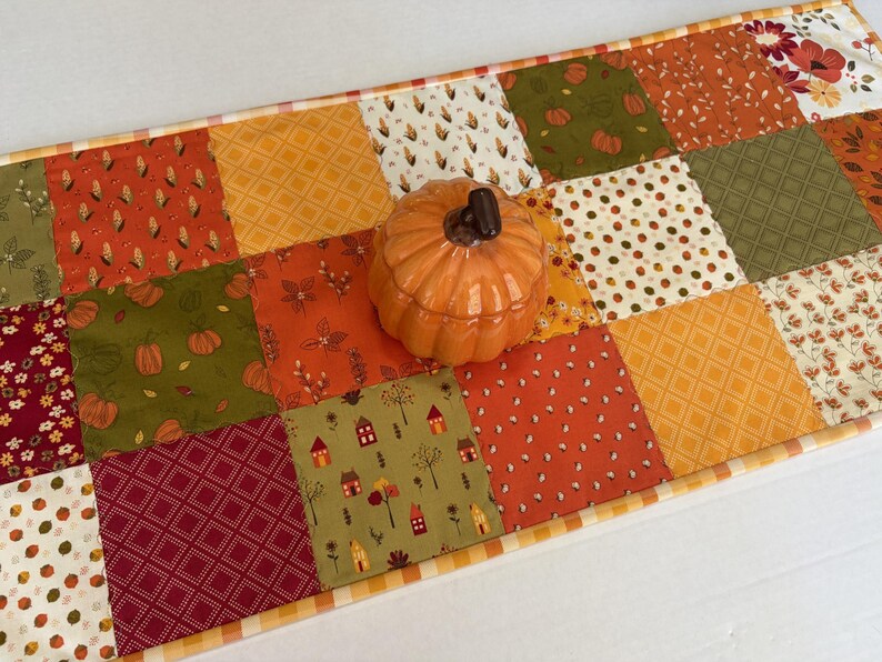 May include: A rectangular patchwork table runner featuring a variety of autumn-themed fabric squares in shades of orange, green, red, and cream, topped with a small, glossy, orange ceramic pumpkin-shaped lidded container in the centre.