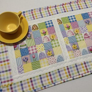 Pode incluir: Uma ch&aacute;vena e um pires amarelos est&atilde;o sobre um caminho de mesa com tema de P&aacute;scoa. O caminho de mesa apresenta um design de patchwork com quadrados pastel, ovos de P&aacute;scoa e padr&otilde;es florais. A borda &eacute; um padr&atilde;o xadrez.