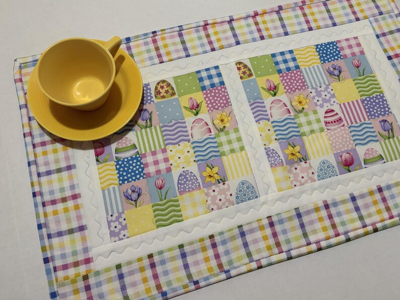 Pode incluir: Uma ch&aacute;vena e um pires amarelos est&atilde;o sobre um caminho de mesa com tema de P&aacute;scoa. O caminho de mesa apresenta um design de patchwork com quadrados pastel, ovos de P&aacute;scoa e padr&otilde;es florais. A borda &eacute; um padr&atilde;o xadrez.