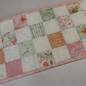 May include: A quilted table runner featuring a patchwork design with various floral and butterfly patterns in shades of pink, peach, and green. The runner has a light pink border and includes inspirational text. The stitches are in a wavy pattern.