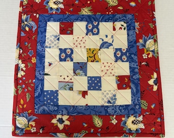 May include: A square quilted fabric piece featuring a central grid of small squares in cream, blue, and red floral patterns. This central section is framed by a solid blue border, which is surrounded by a red outer border with floral motifs.