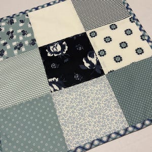 May include: A patchwork quilt square featuring nine fabric squares in shades of blue, white, and cream. The fabrics include floral, geometric, and solid patterns. A blue and white plaid border frames the square.