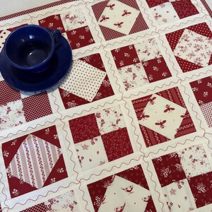 May include: A square patchwork quilt featuring a repeating grid of red and cream fabric squares with various patterns including bees, birds, floral motifs, and polka dots, accented by wavy white stitching and a dark blue ceramic cup and saucer.