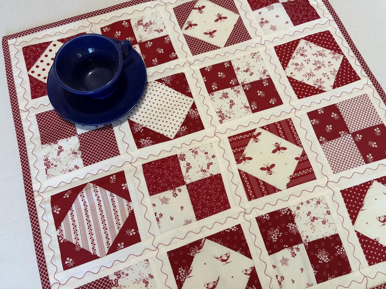 May include: A square patchwork quilt featuring a repeating grid of red and cream fabric squares with various patterns including bees, birds, floral motifs, and polka dots, accented by wavy white stitching and a dark blue ceramic cup and saucer.