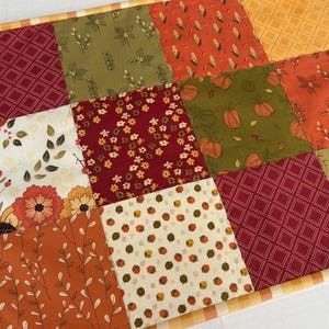 May include: A patchwork quilt featuring square fabric panels with various autumn-themed prints, including pumpkins, corn, flowers, leaves, and small houses in shades of orange, red, green, and cream, finished with a yellow and white chequered border.