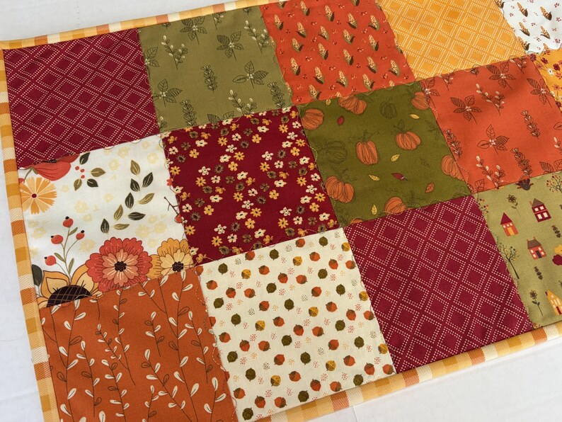 May include: A patchwork quilt featuring square fabric panels with various autumn-themed prints, including pumpkins, corn, flowers, leaves, and small houses in shades of orange, red, green, and cream, finished with a yellow and white chequered border.