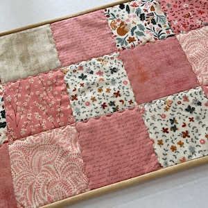 May include: A patchwork placemat with a variety of pink floral and patterned fabrics. The design includes squares with botanical prints, script, and solid pinks, all framed in a light wood border. The overall aesthetic is vintage and feminine.