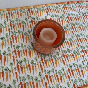 May include: A quilted fabric table runner featuring a repeating pattern of orange carrots with green tops on a white background, bordered by a yellow and white checkered trim, with a cylindrical orange glass candle holder placed on top.