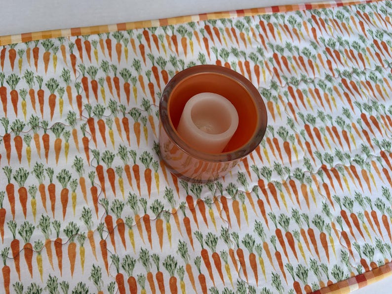 May include: A quilted fabric table runner featuring a repeating pattern of orange carrots with green tops on a white background, bordered by a yellow and white checkered trim, with a cylindrical orange glass candle holder placed on top.