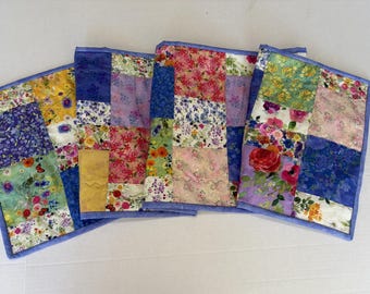 May include: A rectangular quilted table runner featuring a patchwork design of various floral fabrics in shades of blue, pink, yellow, green, and red. The runner is finished with a solid light blue fabric border along all four edges.