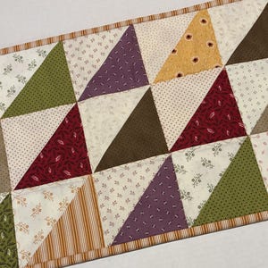 May include: A rectangular table runner featuring a patchwork design of triangles in various colors including red, olive green, purple, brown, and yellow, interspersed with cream-colored fabric squares with small floral and geometric patterns.