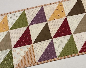 May include: A rectangular table runner featuring a patchwork design of triangles in various colors including red, olive green, purple, brown, and yellow, interspersed with cream-colored fabric squares with small floral and geometric patterns.