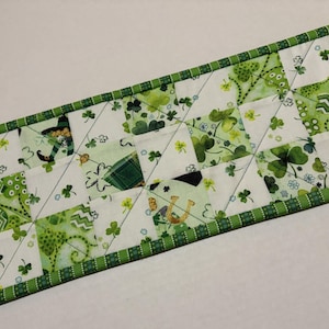 May include: A St. Patrick's Day table runner featuring a patchwork design with green and white squares. The squares are decorated with shamrocks, leprechaun hats, and horseshoes. The edges are trimmed with a green and white striped border.